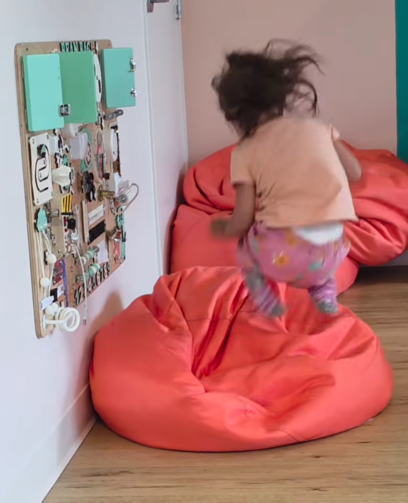 A toddler jumps onto a large bean bag chair in a room with a light wood floor and a white wall decorated with a wooden activity board. Image used as part of the promotion of monthly family memberships at Bean Bag Play Cafe.