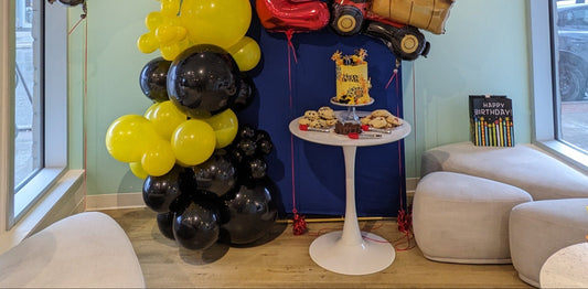 Children's birthday party setup with balloons, a cake, and toys in a room with large windows and comfortable seating.
